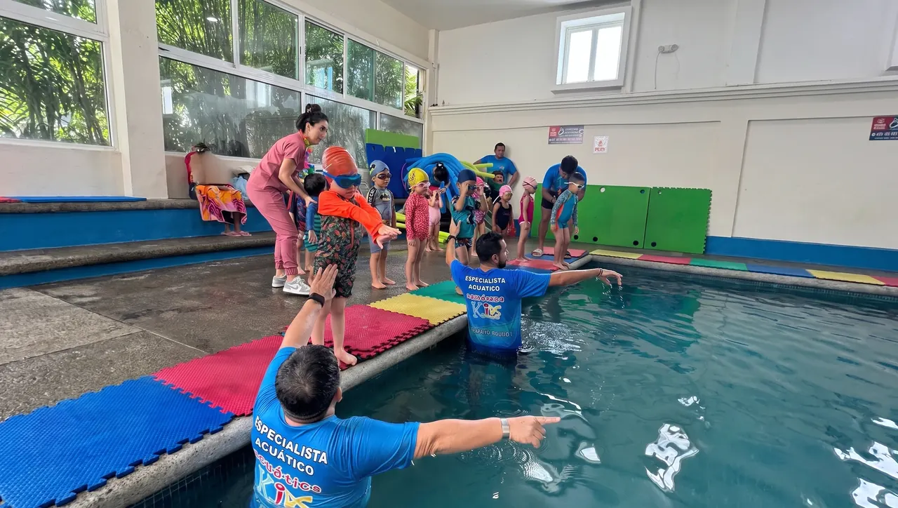 Especialista acuático da indicaciones a niños pequeños en la orilla de la alberca techada de Acuática