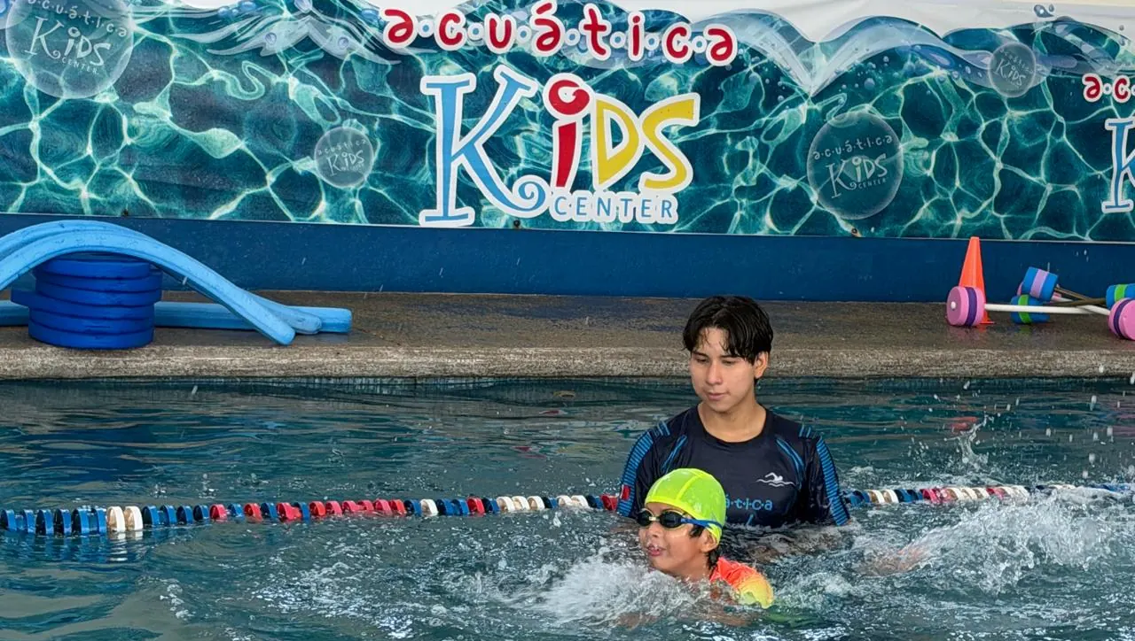 Instructor de Acuática Kids Center acompaña dentro del agua a un niño con goggles durante su clase de natación