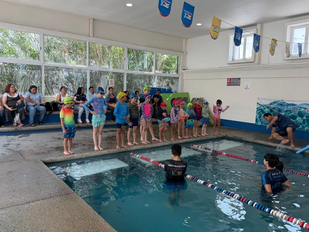 Niñas y niños con gorra y goggles formados en la orilla de la alberca Kids, con instructores en el agua y sus papás mirando la clase
